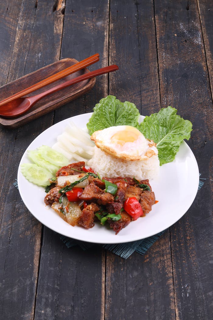A vibrant Thai dish with basil beef, rice, egg, and fresh vegetables on a rustic table.
